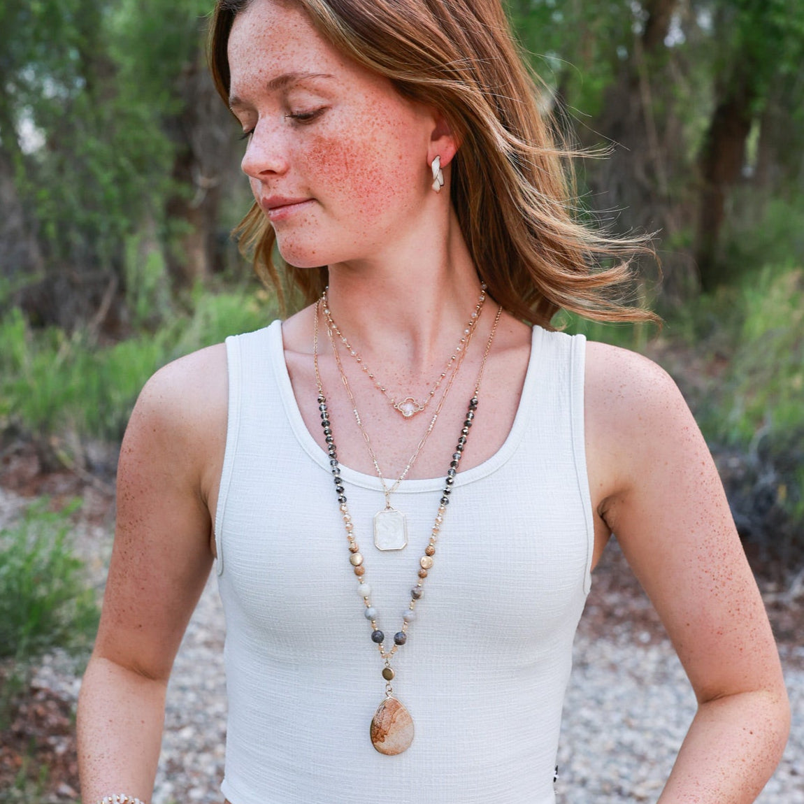 Woman wearing a white tank top and blue jeans with layered necklaces outdoors.